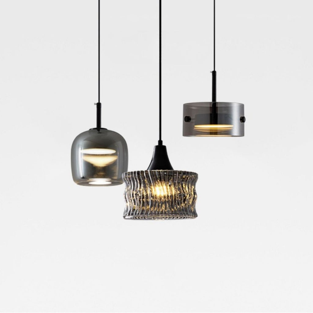 Three modern pendant lights with different glass shades—one rounded, one cylindrical, and one textured—hang from black cords against a plain white background. Two lights are turned on, emitting a warm glow.