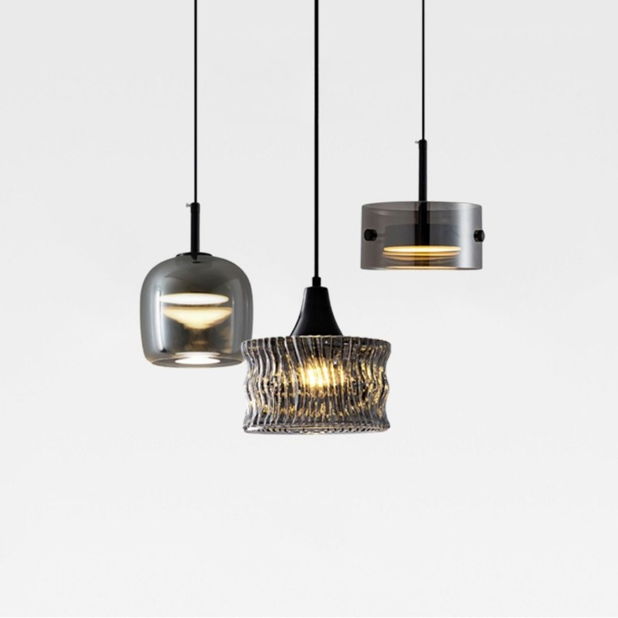 Three modern pendant lights with different glass shades—one rounded, one cylindrical, and one textured—hang from black cords against a plain white background. Two lights are turned on, emitting a warm glow.