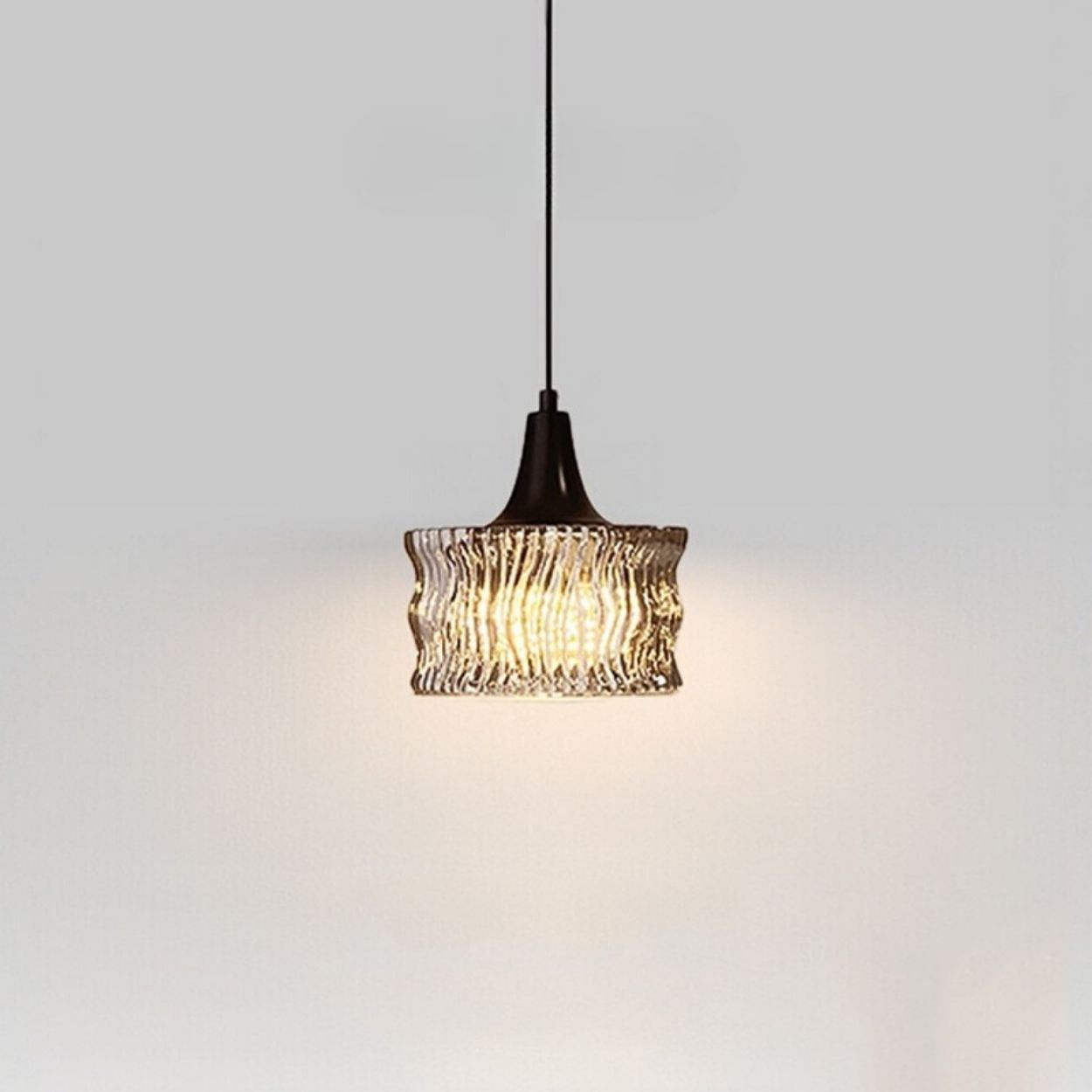 A single pendant light with a black fixture and a wavy, textured glass shade hangs from the ceiling, casting a warm glow on a plain light gray wall.