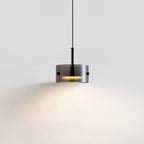 A modern, cylindrical pendant light with a transparent shade hangs from the ceiling, emitting a warm glow and casting soft light on a plain white background.