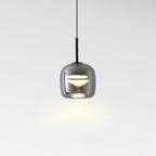 A modern pendant light with a glossy, rounded, transparent gray glass shade hangs from the ceiling, casting a soft glow on a plain white background.