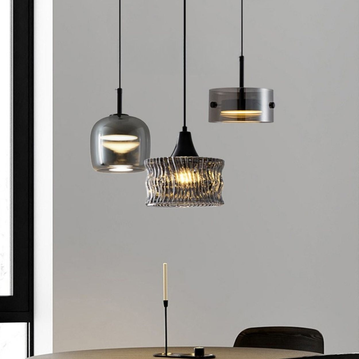 Three modern pendant lights with different glass designs hang above a round table with a single unlit candle in a minimalistic room with neutral colors.
