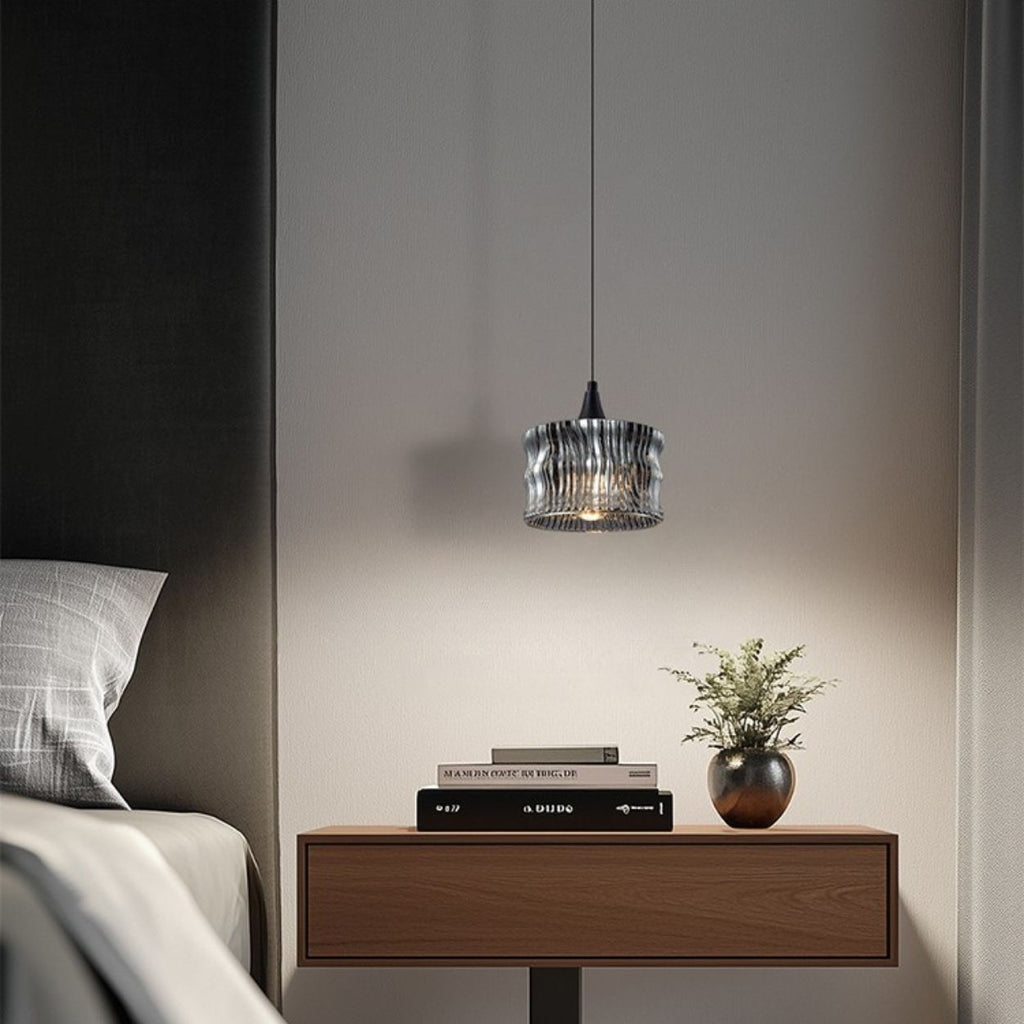 A modern bedroom scene with a wooden nightstand, stacked books, a small potted plant, and a pendant light hanging above, next to a bed with gray and white bedding.