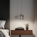 A modern bedroom scene with a wooden nightstand, stacked books, a small potted plant, and a pendant light hanging above, next to a bed with gray and white bedding.
