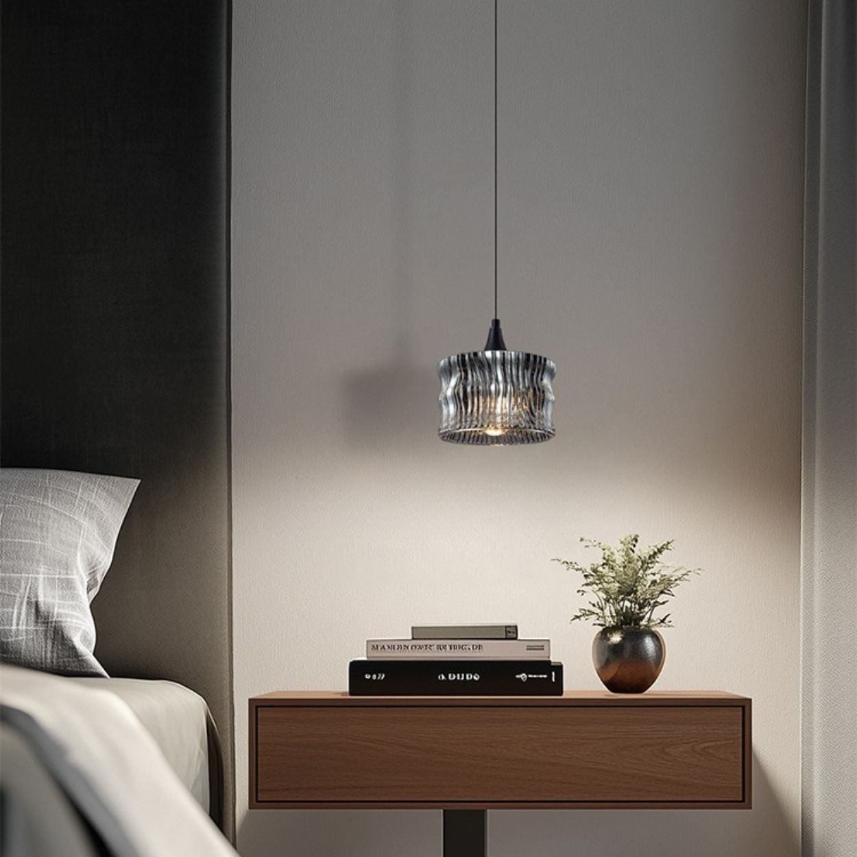A modern bedroom scene with a wooden nightstand, stacked books, a small potted plant, and a pendant light hanging above, next to a bed with gray and white bedding.