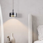 A modern bedroom corner with a plush light-gray headboard, white pillows, a round white table, minimalist vases, and a sleek black and silver pendant light casting a warm glow on the wall.