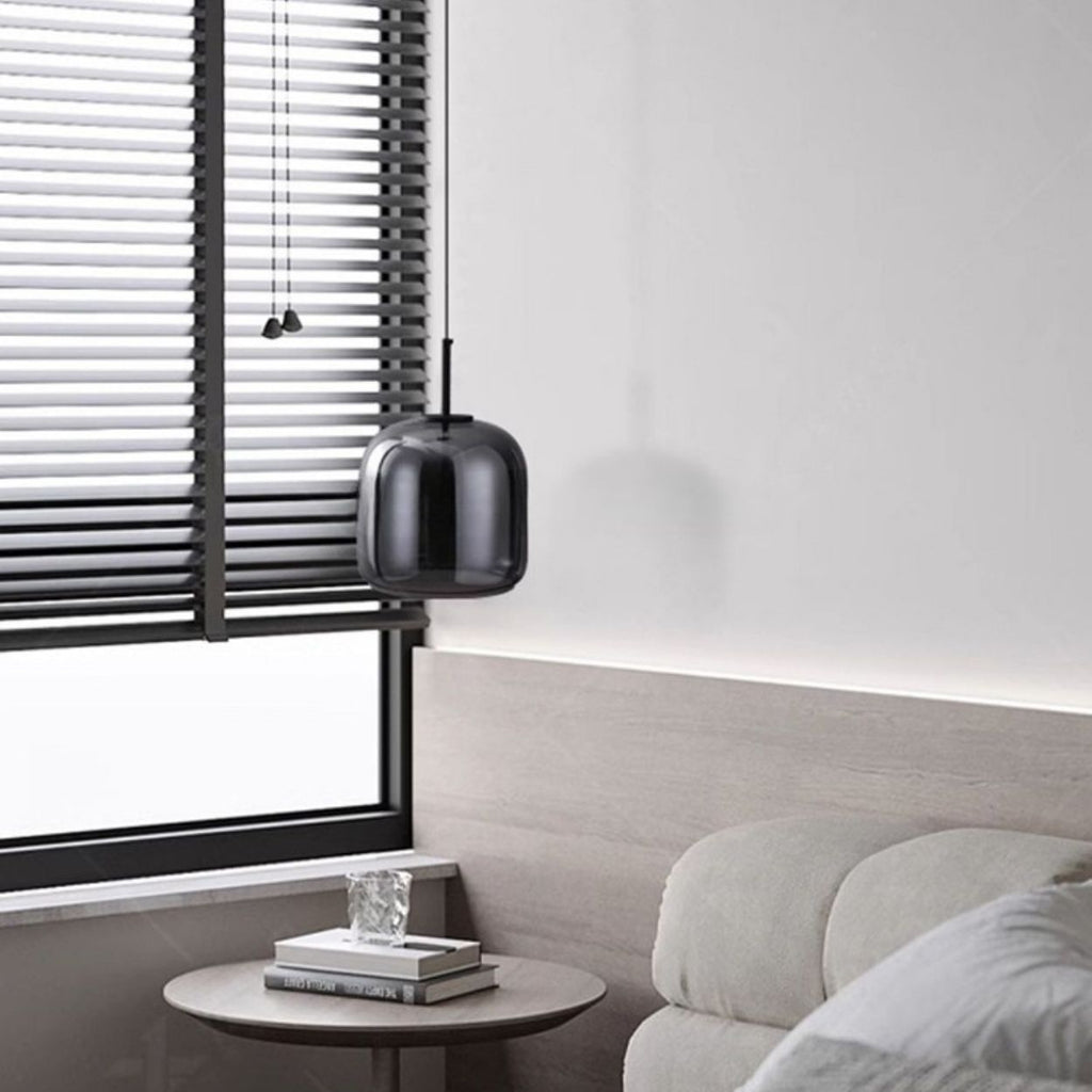 A modern, minimalistic living room corner with a light gray sofa, a small round side table holding two books and a glass ornament, a black pendant lamp, and horizontal black blinds covering a large window.