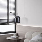 A modern, minimalistic living room corner with a light gray sofa, a small round side table holding two books and a glass ornament, a black pendant lamp, and horizontal black blinds covering a large window.