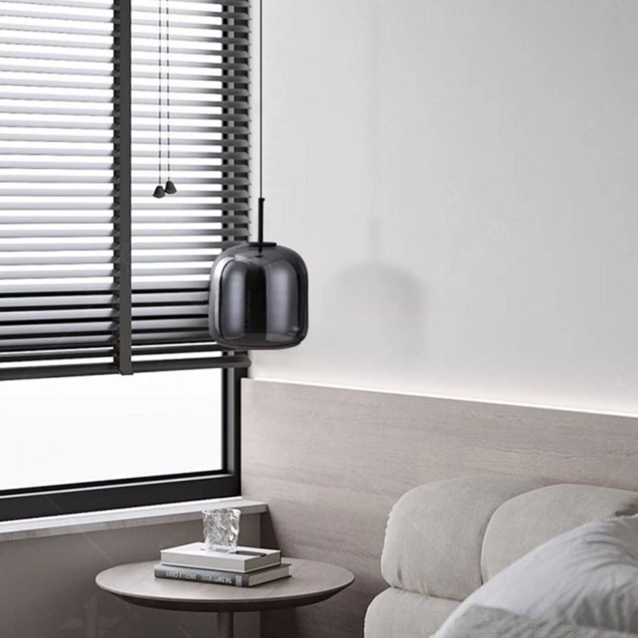A modern, minimalistic living room corner with a light gray sofa, a small round side table holding two books and a glass ornament, a black pendant lamp, and horizontal black blinds covering a large window.