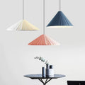 Three cone-shaped pendant lights in white, peach, and blue hang above a small round table with minimalist vases and a sprig of greenery, set against a light gray wall.