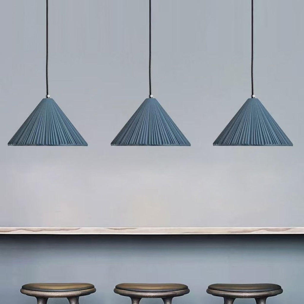 Three blue, pleated pendant lights hang from the ceiling above a light-colored countertop, with three dark, round stools positioned below the counter against a light gray wall.