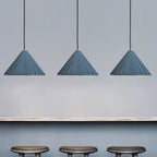 Three blue, pleated pendant lights hang from the ceiling above a light-colored countertop, with three dark, round stools positioned below the counter against a light gray wall.