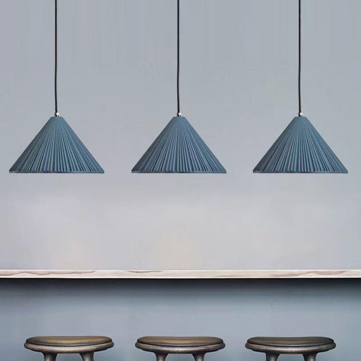 Three blue, pleated pendant lights hang from the ceiling above a light-colored countertop, with three dark, round stools positioned below the counter against a light gray wall.