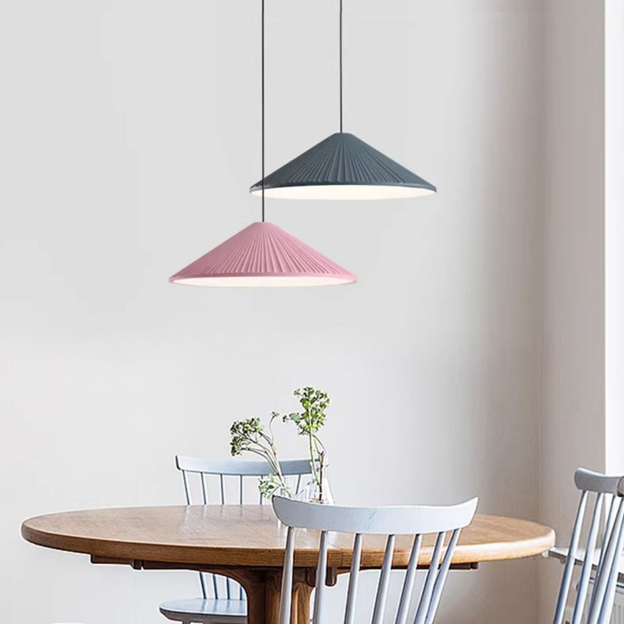 A round wooden table with light gray chairs, a small vase with green plants, and two stylish pendant lights—one pink and one dark gray—hanging above against a light-colored wall.