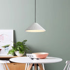 A modern dining area with a round white table, a pink-striped bowl, a white chair, a green potted fern, a framed photo, and a striking pendant light hanging against the muted green wall.