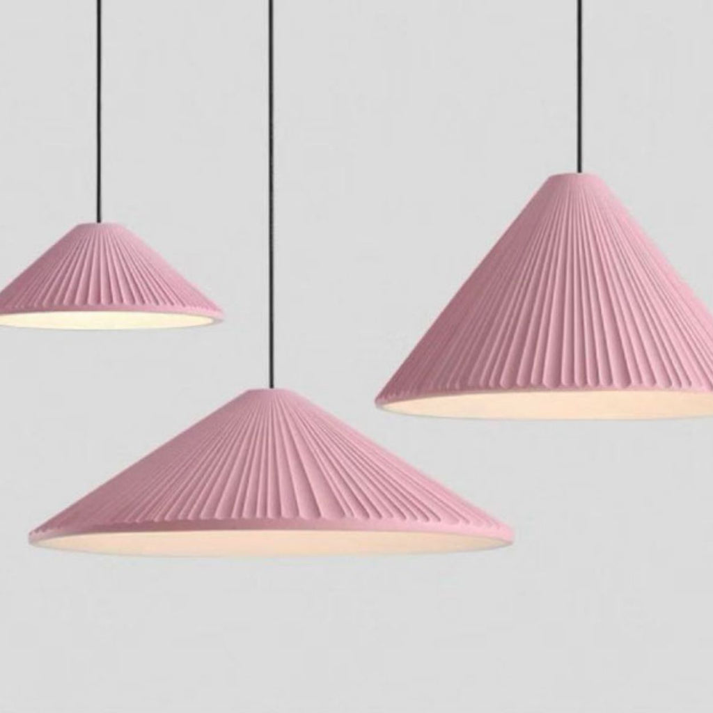 Three pendant lights with pleated, cone-shaped pink lampshades hang from black cords against a light gray background. Each Pendant Light features a ribbed texture and emits a soft white glow underneath.