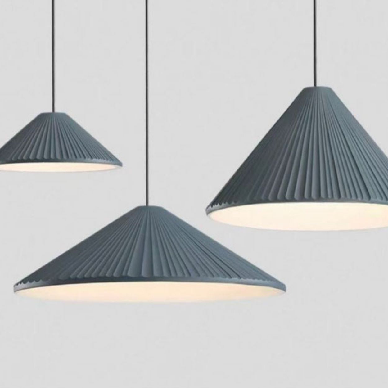 Three hanging pendant lights with ribbed, cone-shaped gray shades, suspended at different heights against a plain light gray background. The lamps are lit, casting a soft glow underneath.