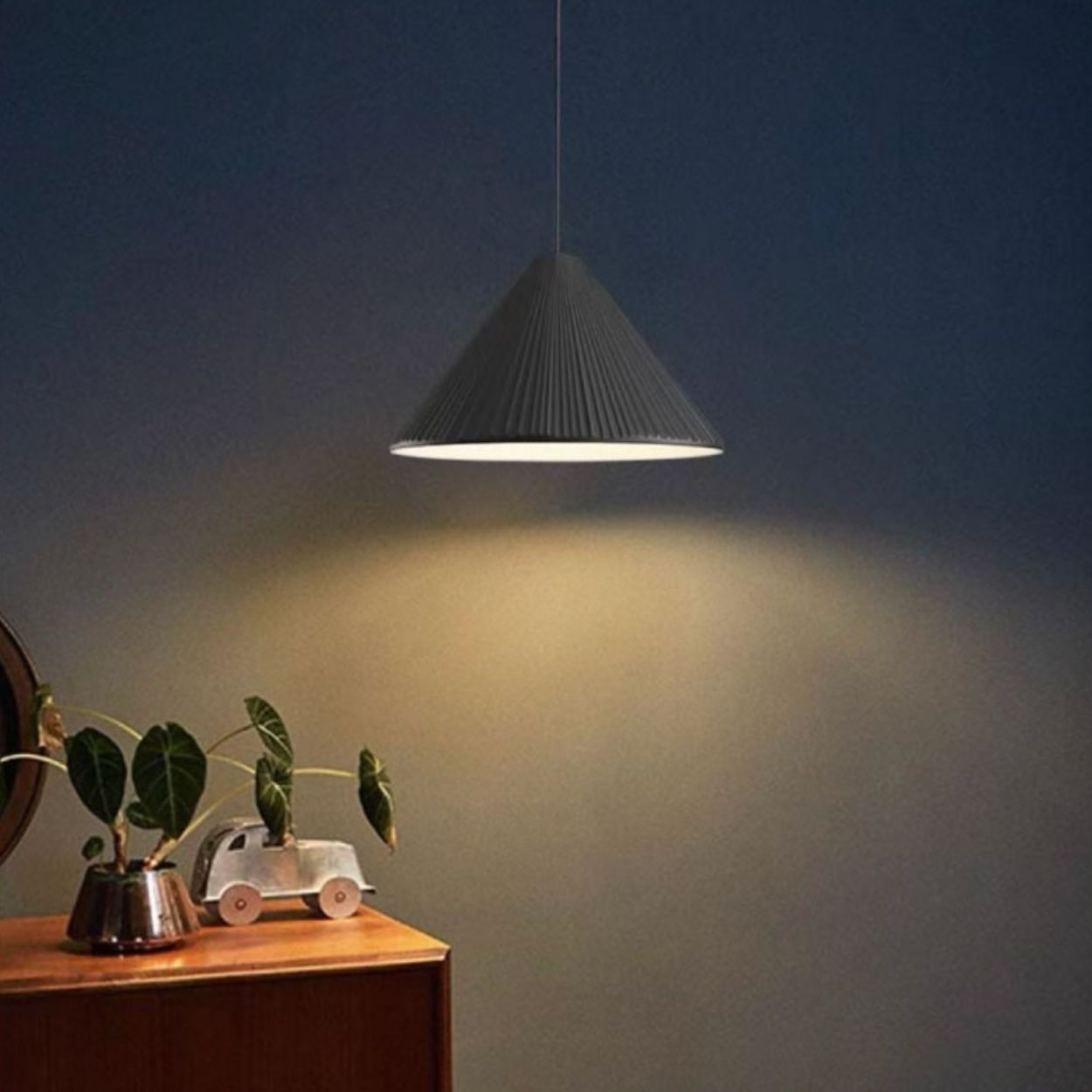 A cone-shaped black pendant light hangs above a wooden table with a small potted plant and a toy car, casting warm light on a dark blue wall.