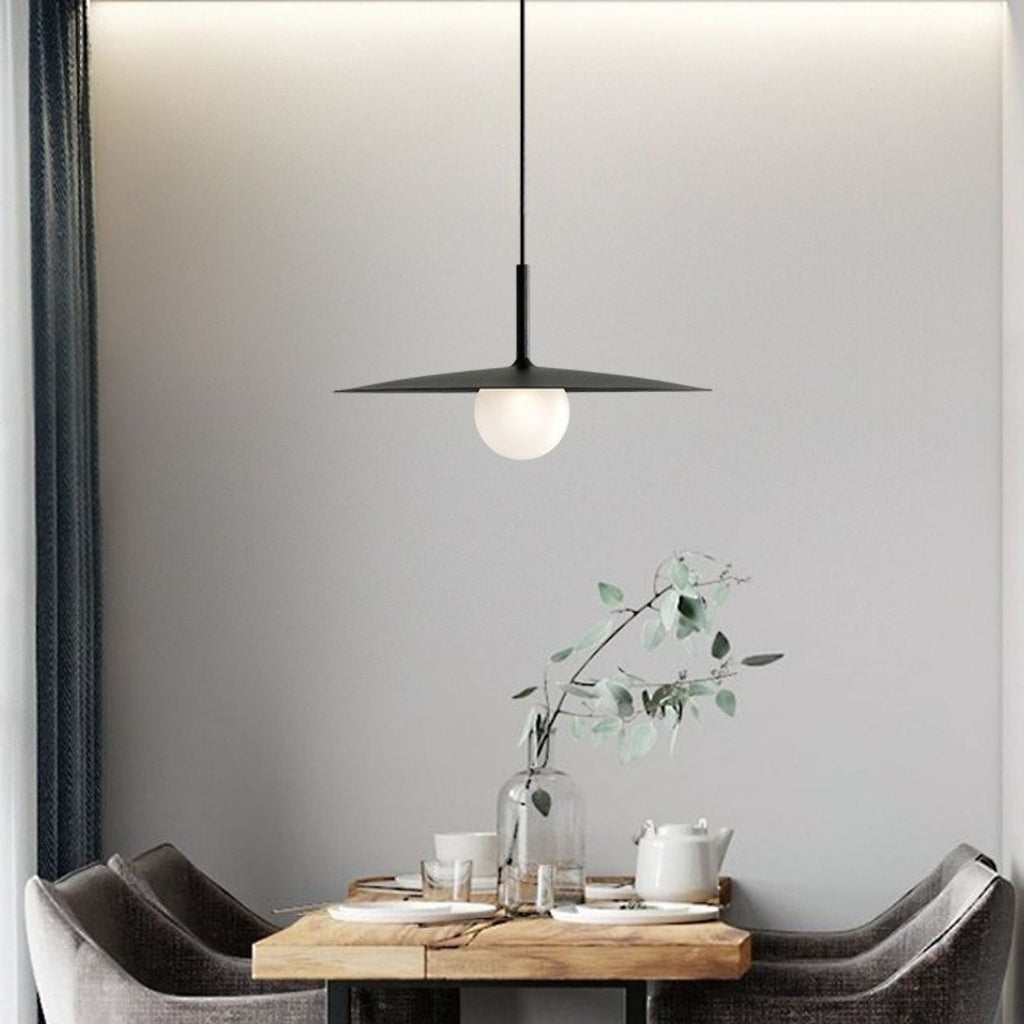 A modern dining area features a wooden table set for tea, two gray chairs, a glass vase with eucalyptus branches, and a striking pendant lamp hanging above against a light gray wall.