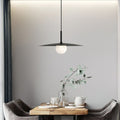 A modern dining area features a wooden table set for tea, two gray chairs, a glass vase with eucalyptus branches, and a striking pendant lamp hanging above against a light gray wall.