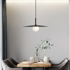 A modern dining area features a wooden table set for tea, two gray chairs, a glass vase with eucalyptus branches, and a striking pendant lamp hanging above against a light gray wall.