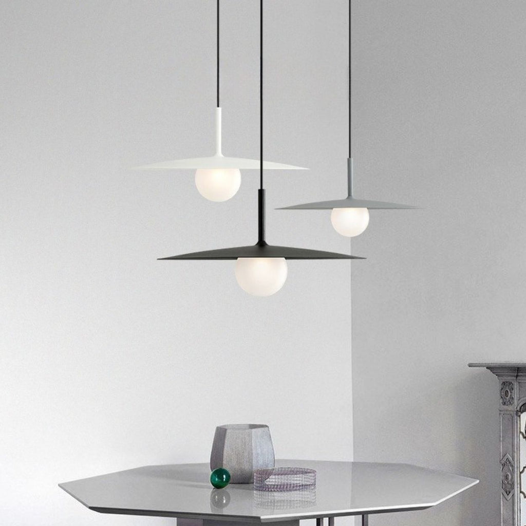 Three modern pendant lamps with flat, circular shades hang above a sleek, hexagonal table. A small decorative object and a glass ornament rest on the table against a minimalist, light gray background.