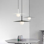 Three modern pendant lamps with flat, circular shades hang above a sleek, hexagonal table. A small decorative object and a glass ornament rest on the table against a minimalist, light gray background.