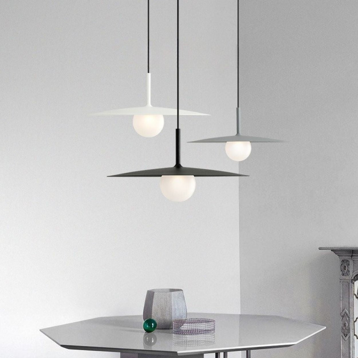 Three modern pendant lamps with flat, circular shades hang above a sleek, hexagonal table. A small decorative object and a glass ornament rest on the table against a minimalist, light gray background.