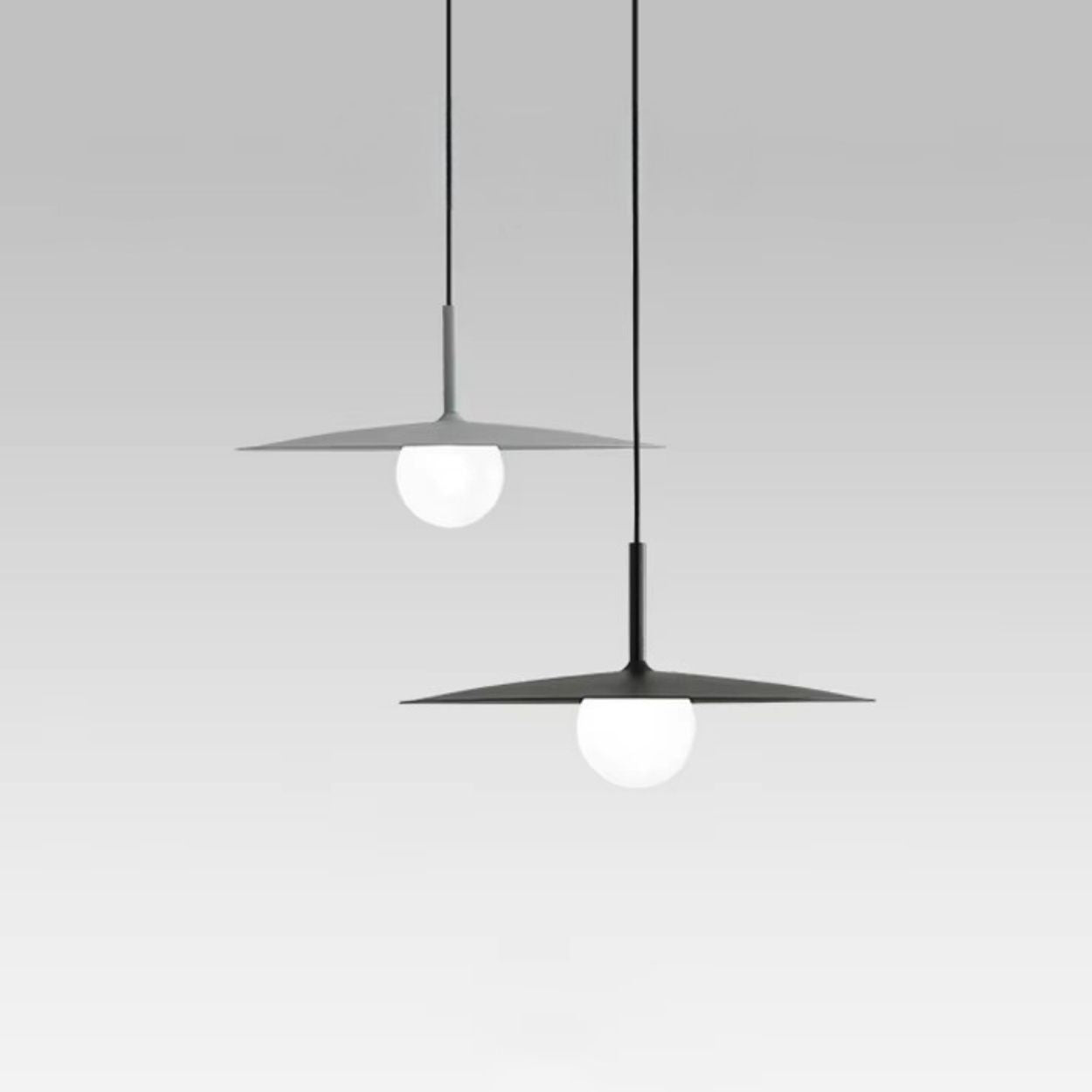 Two modern pendant lights hang from the ceiling, each featuring a flat, circular shade with a round white bulb beneath, set against a plain light gray background.