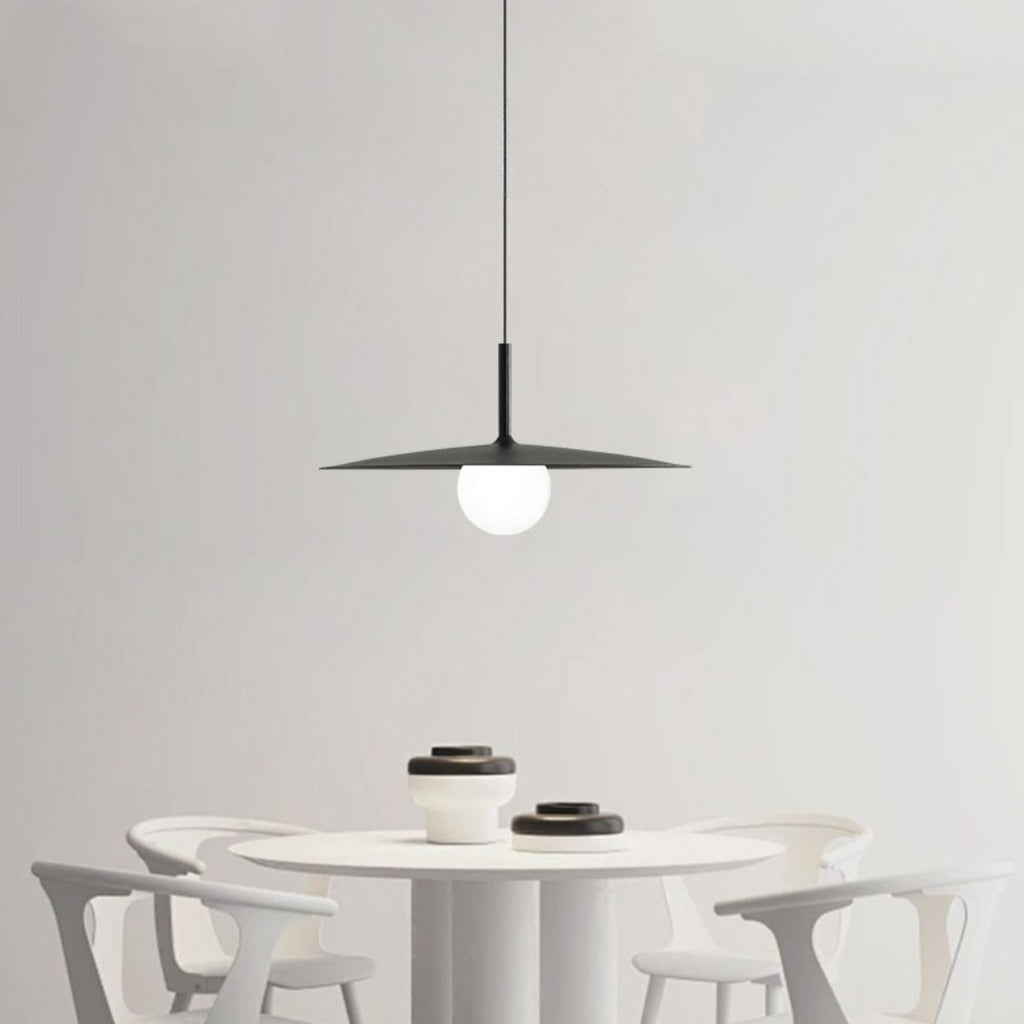 A modern dining area with a round white table, four white chairs, and black-and-white bowls. A black pendant light with a round bulb hangs above the table against a plain white wall.