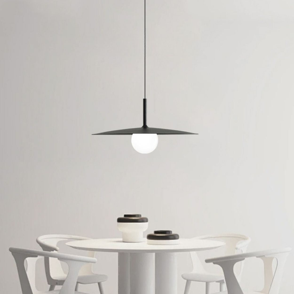 A modern dining area with a round white table, four white chairs, and black-and-white bowls. A black pendant light with a round bulb hangs above the table against a plain white wall.