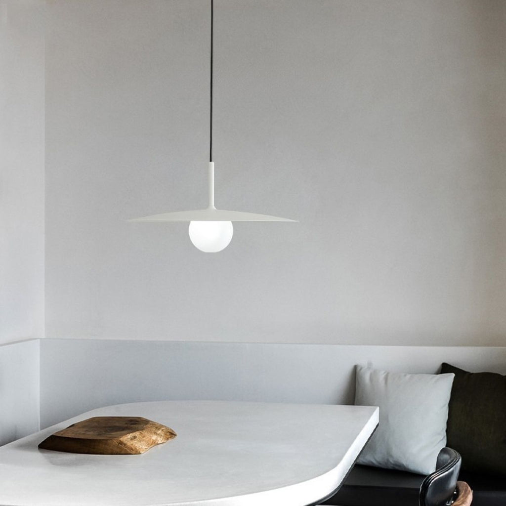 A minimalist dining area with a white curved table, a wooden serving board, a black chair, gray and white cushions, and a modern, white pendant light hanging above. The walls are light gray and unadorned.