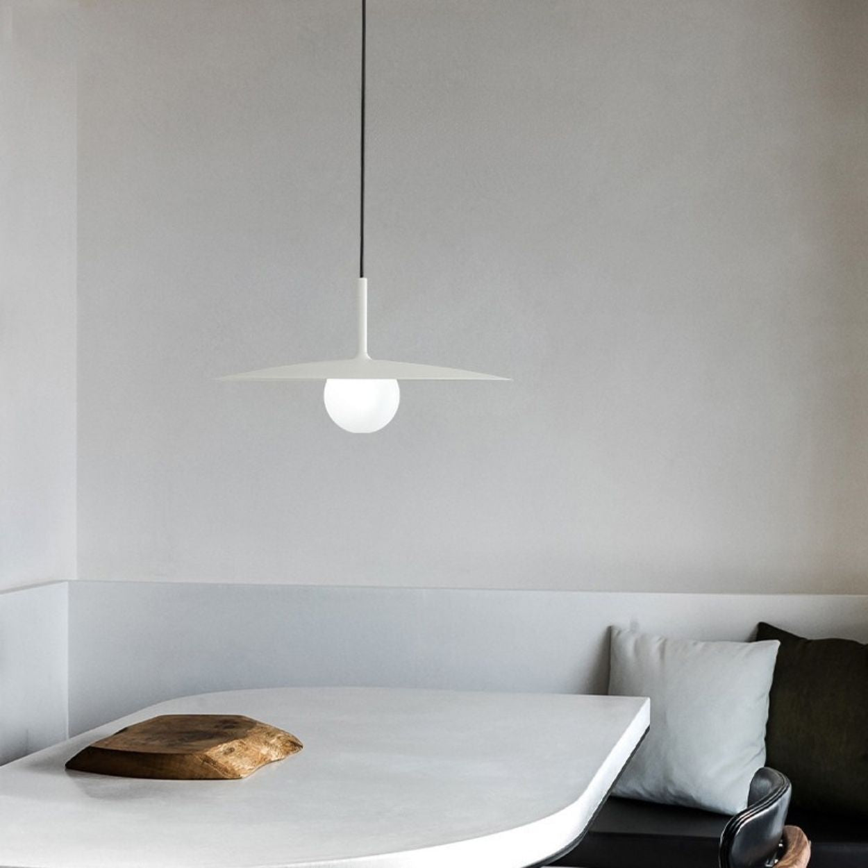 A minimalist dining area with a white curved table, a wooden serving board, a black chair, gray and white cushions, and a modern, white pendant light hanging above. The walls are light gray and unadorned.