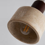 A close-up of a hanging pendant light fixture with a cylindrical, textured beige stone lampshade and an exposed bulb inside. The fixture is suspended from above against a plain light gray background.
