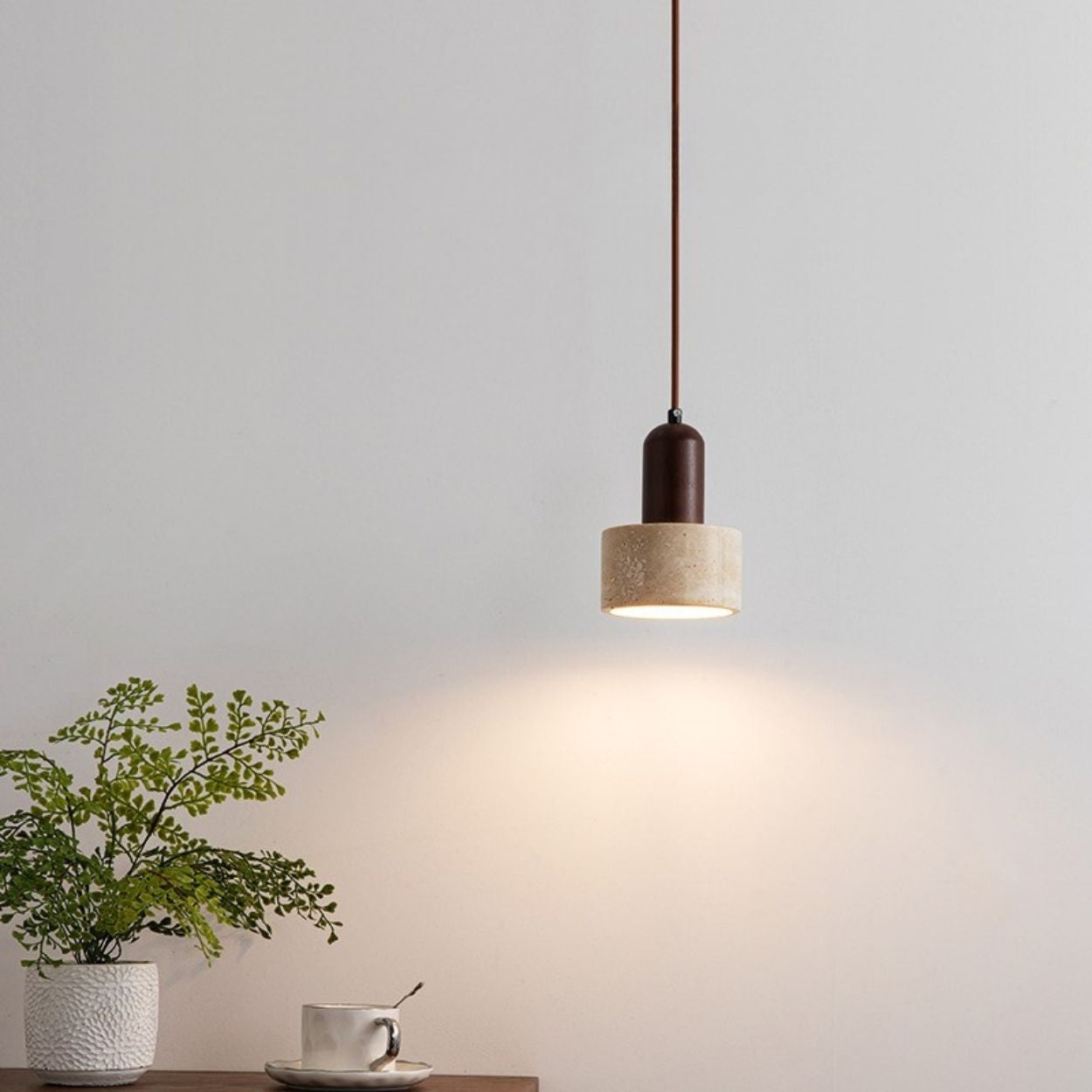 A modern pendant light hangs from the ceiling, illuminating a plain white wall. Below, a wooden table holds a white textured plant pot with greenery and a white cup and saucer, all gently lit by the stylish pendant light above.