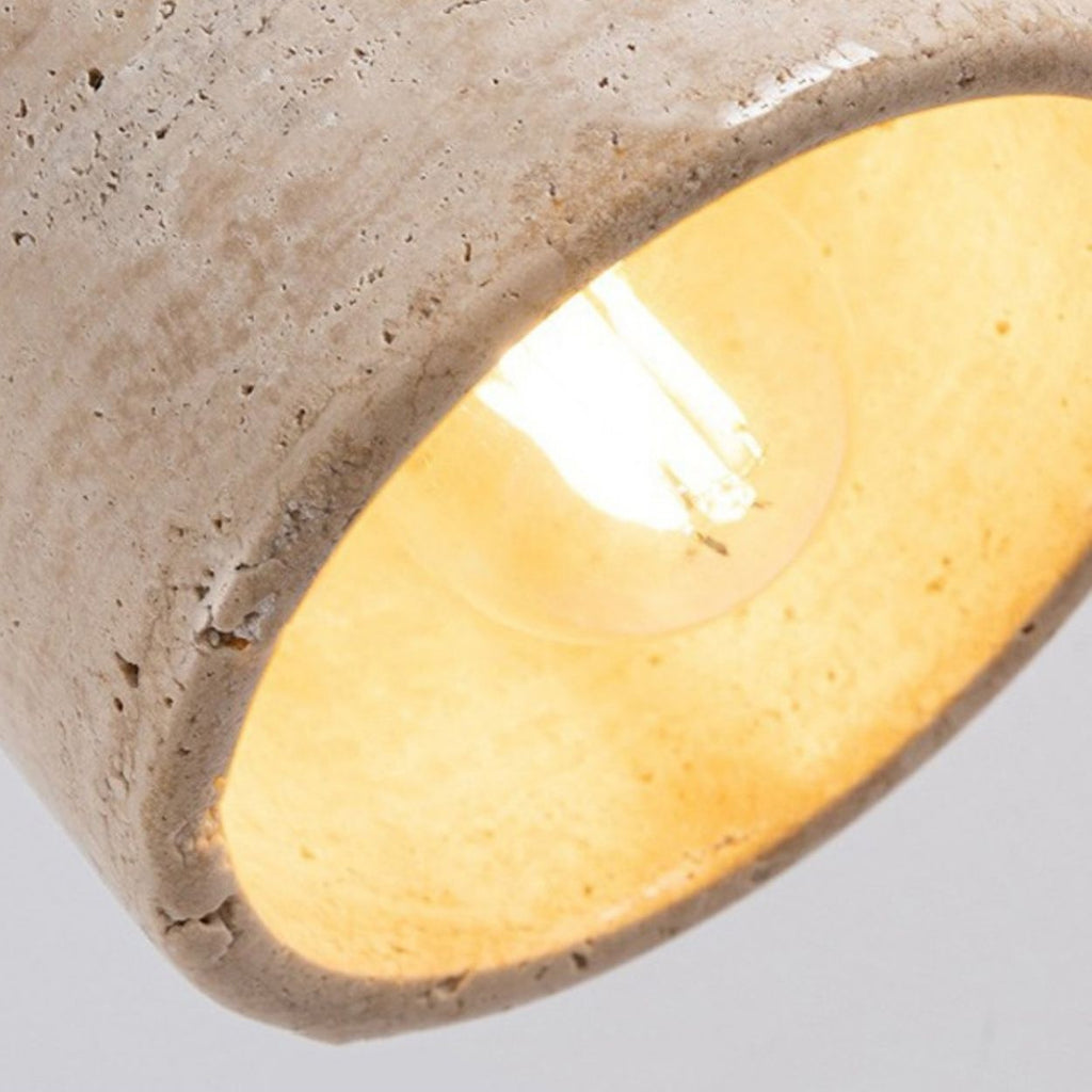Close-up of a glowing light bulb inside a textured, beige concrete pendant light. The rough surface of the lampshade contrasts with the warm, soft light emitted from the bulb.
