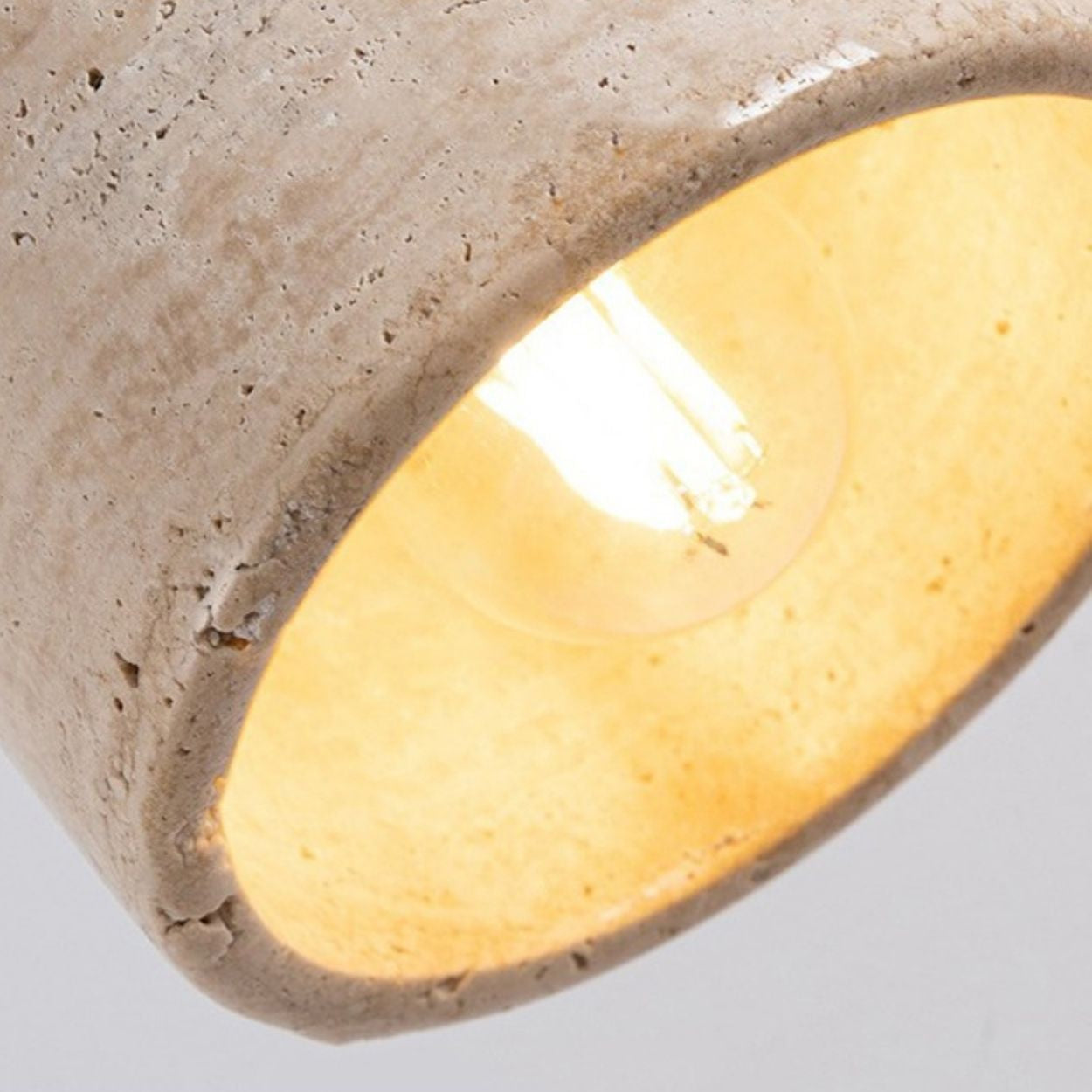 Close-up of a glowing light bulb inside a textured, beige concrete pendant light. The rough surface of the lampshade contrasts with the warm, soft light emitted from the bulb.