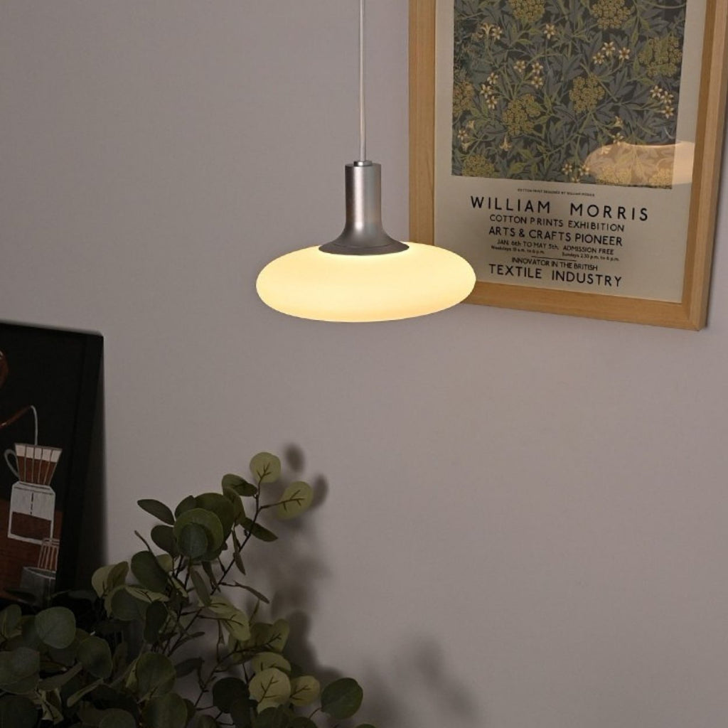 A modern pendant lamp with a glowing white shade hangs from the ceiling near a framed William Morris exhibition poster and a leafy green plant.