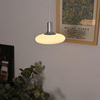 A modern pendant lamp with a glowing white shade hangs from the ceiling near a framed William Morris exhibition poster and a leafy green plant.