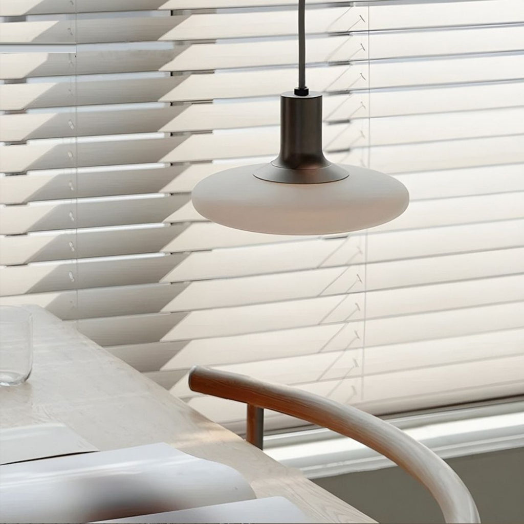 A modern pendant lamp hangs above a table set with placemats and a glass, next to a wooden chair and large window with white blinds. Sunlight softly filters through the blinds.
