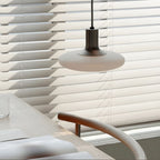 A modern pendant lamp hangs above a table set with placemats and a glass, next to a wooden chair and large window with white blinds. Sunlight softly filters through the blinds.