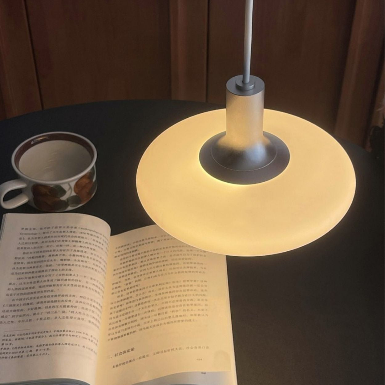 A glowing pendant lamp hangs over an open book written in Chinese on a dark table, with a cup of coffee or tea placed nearby.