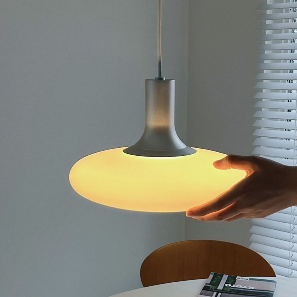 A hand gently touches a glowing, modern pendant light suspended above a round table near window blinds. A chair and a book or magazine are partially visible on the table.