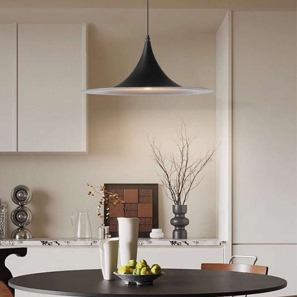 A modern dining room features a round table with green apples, two white vases, and a cup. A striking black pendant light hangs above. In the background, white cabinets, a marble countertop, and a decorative vase with branches complete the look.