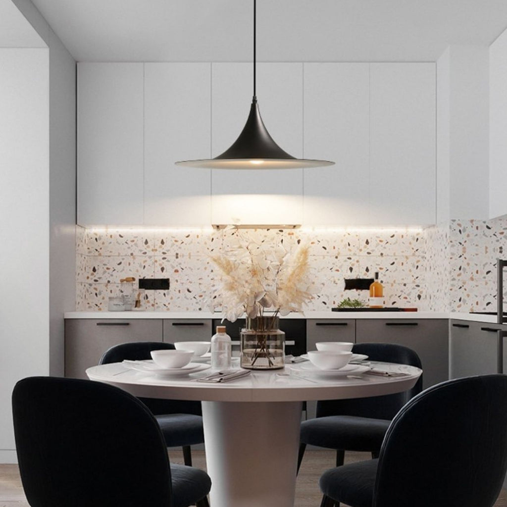 A modern kitchen with white cabinets and a terrazzo backsplash features a round dining table for four with black chairs. A striking pendant light hangs above the table, highlighting the dried flowers used as a centerpiece.