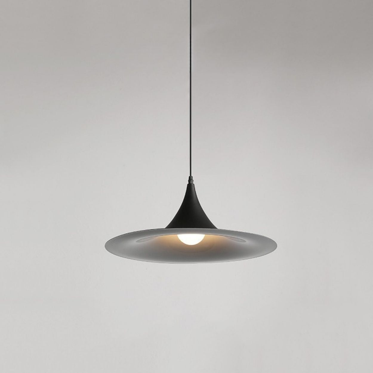 A modern, black pendant lamp with a wide, shallow shade hangs from the ceiling, emitting a soft light against a plain, light gray background.