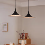 Two modern black pendant lights hang above a wooden dining table with a white vase holding branches. Minimalist decor, with framed wall art and decorative letters in the background. Neutral, softly lit room.