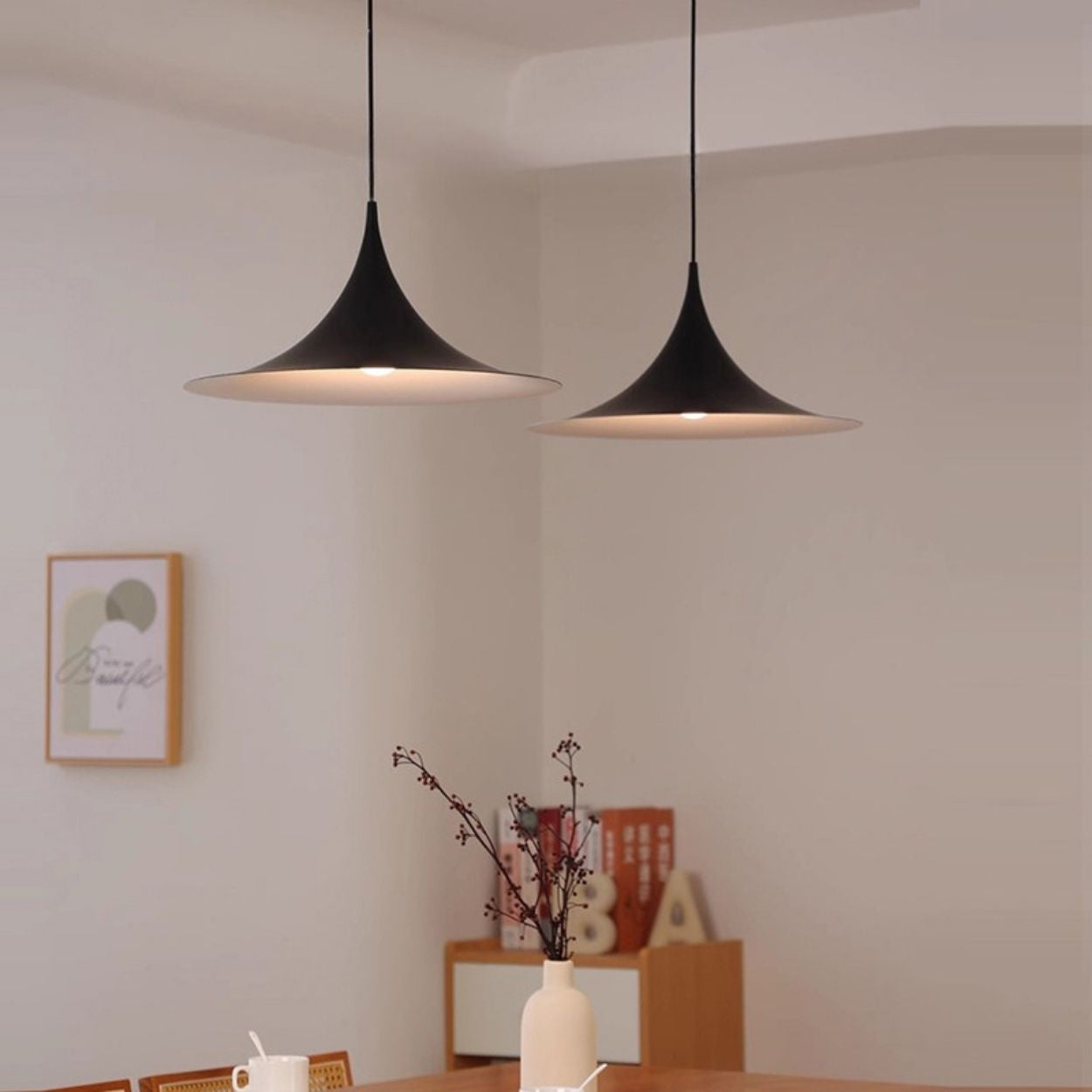 Two modern black pendant lights hang above a wooden dining table with a white vase holding branches. Minimalist decor, with framed wall art and decorative letters in the background. Neutral, softly lit room.