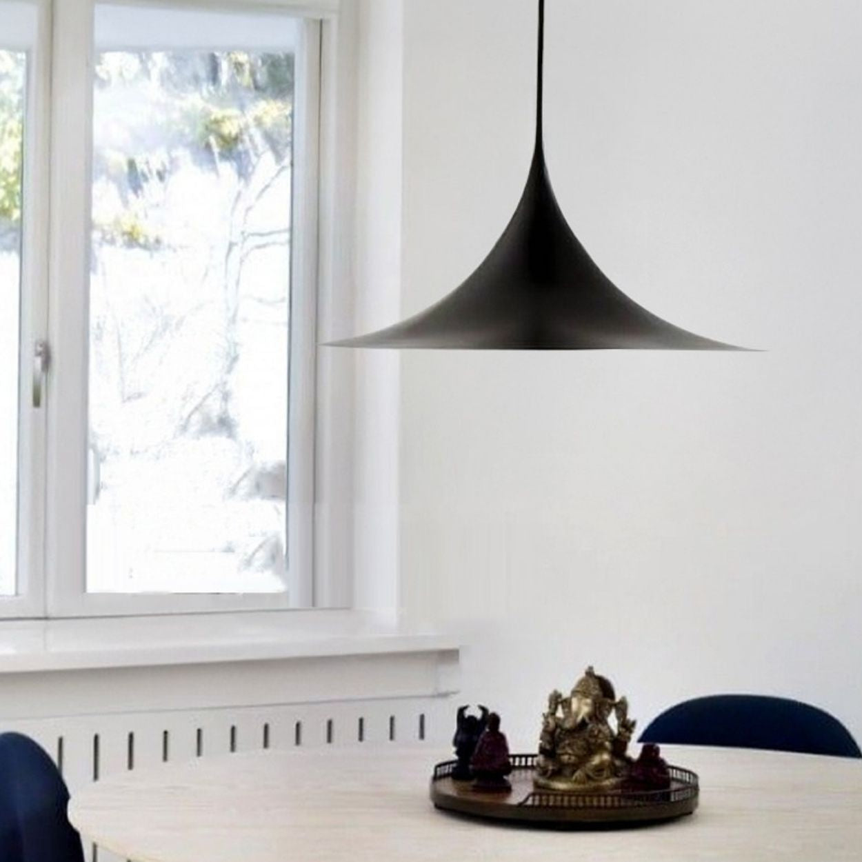 A minimalist dining area with a round table, blue chairs, a tray holding a small Ganesha statue, and a sleek Pendant Light above, set near a window with white walls and natural light.