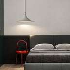 A modern bedroom features a gray upholstered bed, two pillows, a gray blanket, and a red side table with a glass. A sleek pendant light hangs above, casting warm illumination on the wall for a cozy atmosphere.